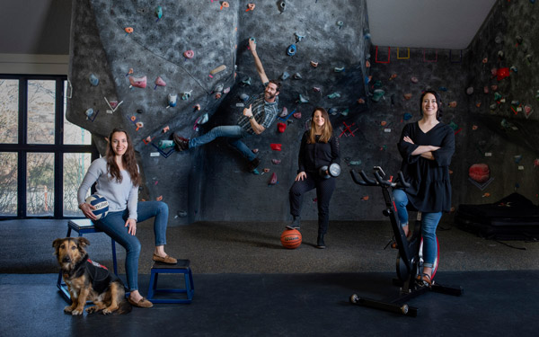 Four people pose in a gym with a rock climbing wall, a service dog, and sports equipment, representing different activities.