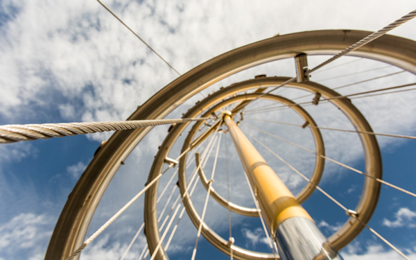 Upward view of a metal sculpture with circular rings and cables against a bright blue sky with clouds on the Regis campus.