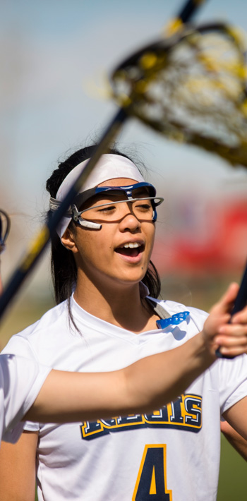 Regis University women’s lacrosse players cheer with raised sticks, wearing white jerseys and protective eyewear.