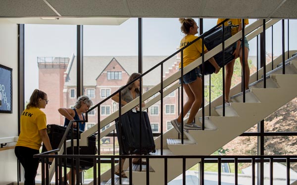 Students and family members carry luggage up a dormitory staircase during move-in day, assisted by volunteers in yellow shirts.