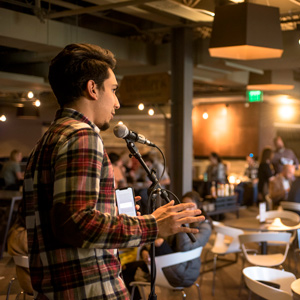 A student speaks into a microphone at Walker’s Pub, addressing a lively audience seated at tables under warm lighting.