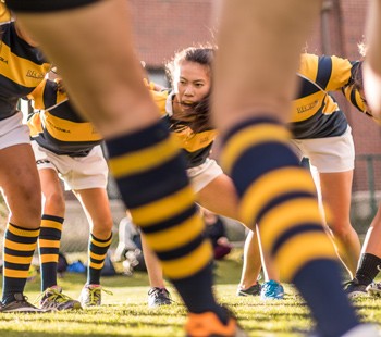 Regis University women’s rugby team prepares for a scrum, wearing navy and gold jerseys, striped socks, and mouthguards.