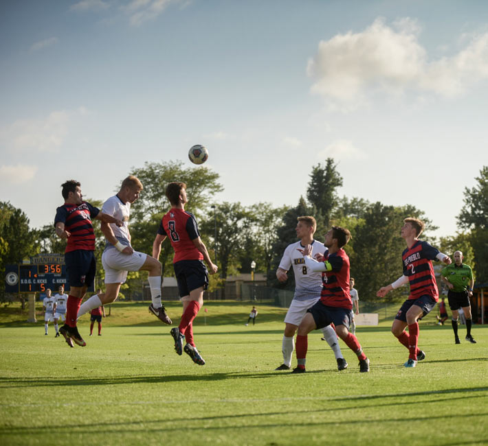 Regis University men’s soccer players in white and opponents in red and navy compete for a header during a match on campus.
