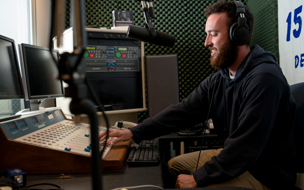 A student wearing headphones adjusts a soundboard in a radio station, speaking into a microphone in a soundproofed room.