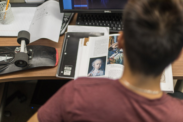 Student studying an art history book at a desk with a skateboard, notebooks, and a computer in a workspace setting.