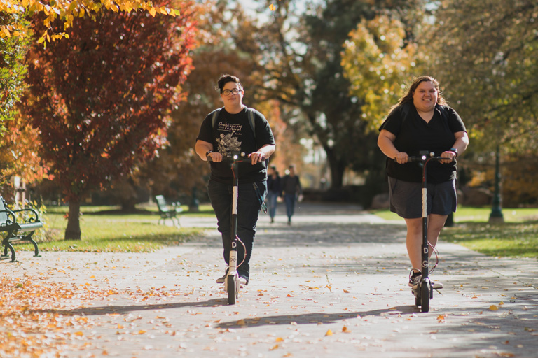 two students ride electric scooters on a tree-lined campus path in autumn