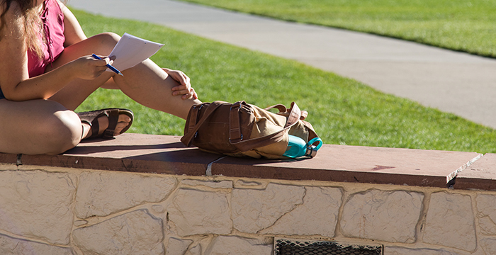 student sitting on a wall beside her backpack doing homework 