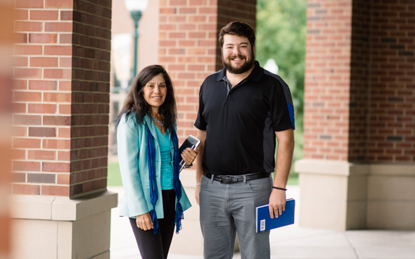 Smiling adults stand outside a brick building on campus, holding books and a phone.