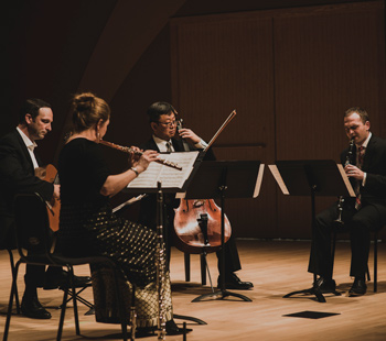 Musicians performing a chamber music piece with guitar, flute, cello and clarinet in a concert setting.