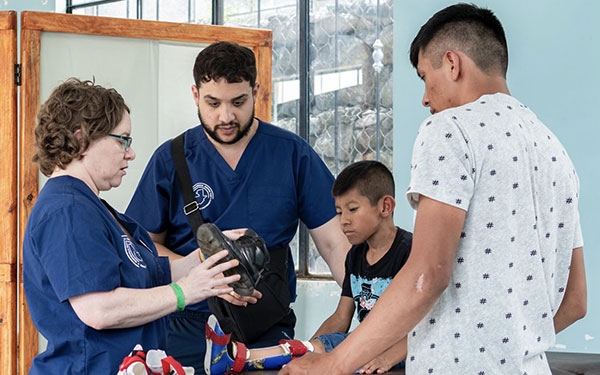 students fit a shoe on a small boy seated on an exam table while his guardian stands nearby