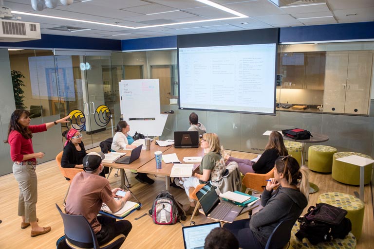 Instructor presenting to a small class of students in a collaborative learning environment. The students are using laptops and reference materials while viewing a presentation on a large screen.