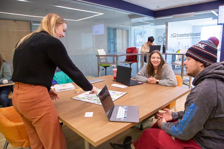 A student standing at a wooden table points to school work while two other students sit and engage in a discussion within a modern glass walled classroom.