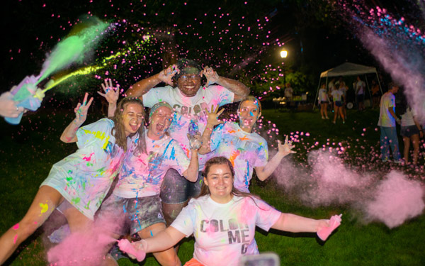 Students at Regis University celebrate a nighttime color run event, covered in bright paint and throwing colorful powder in the air