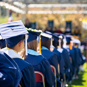 a row of graduates wearing caps and gowns watches their commencement ceremony on the Regis campus