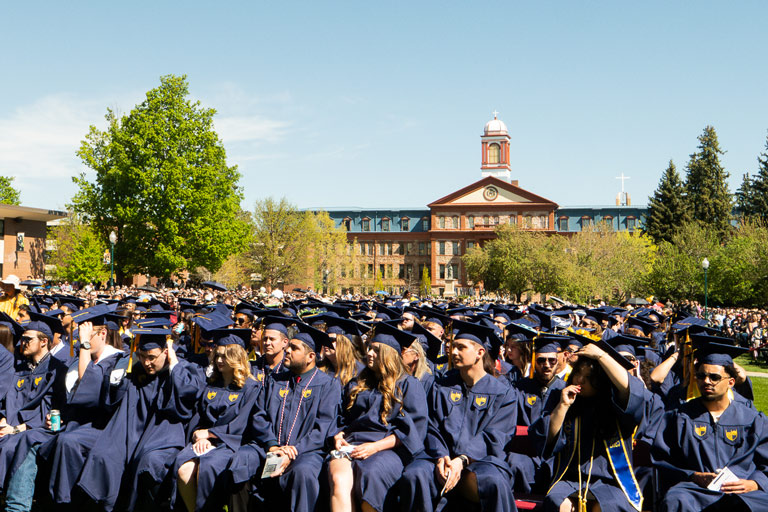 A crowd of graduates in gowns and caps seated outdoors during graduation ceremony, with Main Hall and trees in the background