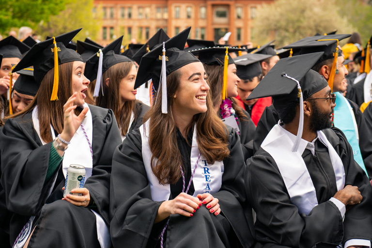 Graduates in black caps and gowns sit together outdoors, smiling and chatting during a commencement ceremony.