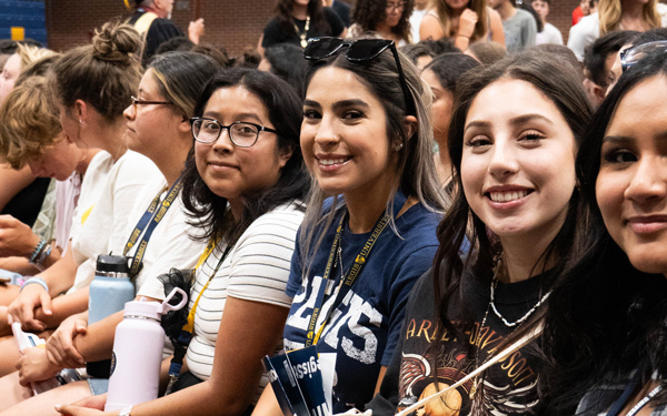 Students sit together in a gymnasium, smiling and holding school gear during an orientation event.