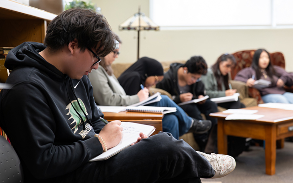 A group of six students sitting in a cozy room, focused on writing in notebooks