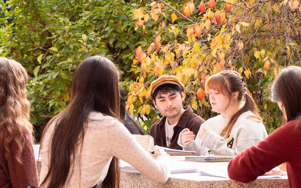 A group of students sits outdoors at a table, engaged in discussion, surrounded by vibrant fall foliage
