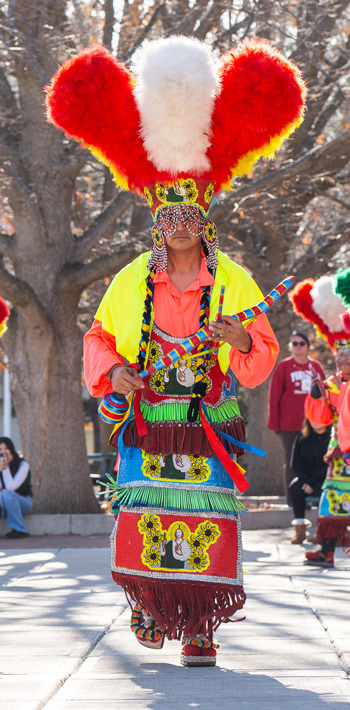 Dancers in colorful traditional attire with tall feathered headdresses perform Danza de Matachines in an outdoor procession.