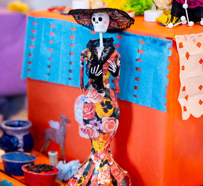 A figurine of La Catrina in a floral dress and hat stands on a colorful altar decorated with papel picado and various small objects