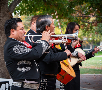 Four mariachi musicians perform outside, wearing traditional black outfits with silver embroidery, playing trumpet, violin, and guitar