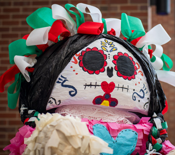 Colorful Día de los Muertos piñata with a white skull face, black eyes, red flowers, heart designs, and green, white, and red ribbon hair against a brick background