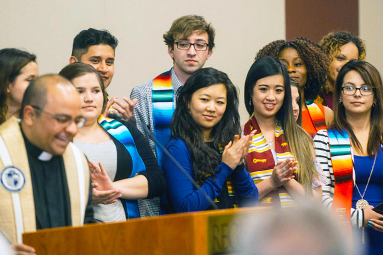 a priest speaks before a group of students wearing diversity stoles at the diversity ceremony during commencement