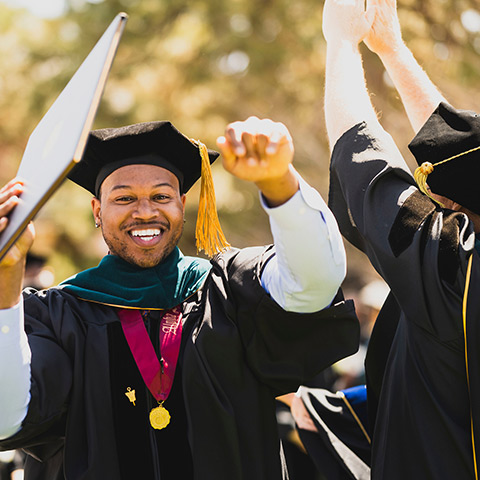 a doctoral graduate wearing regalia smiles and raises his hands in celebration as faculty cheer at the Spring commencement ceremony on the Quad on the Northwest Denver campus