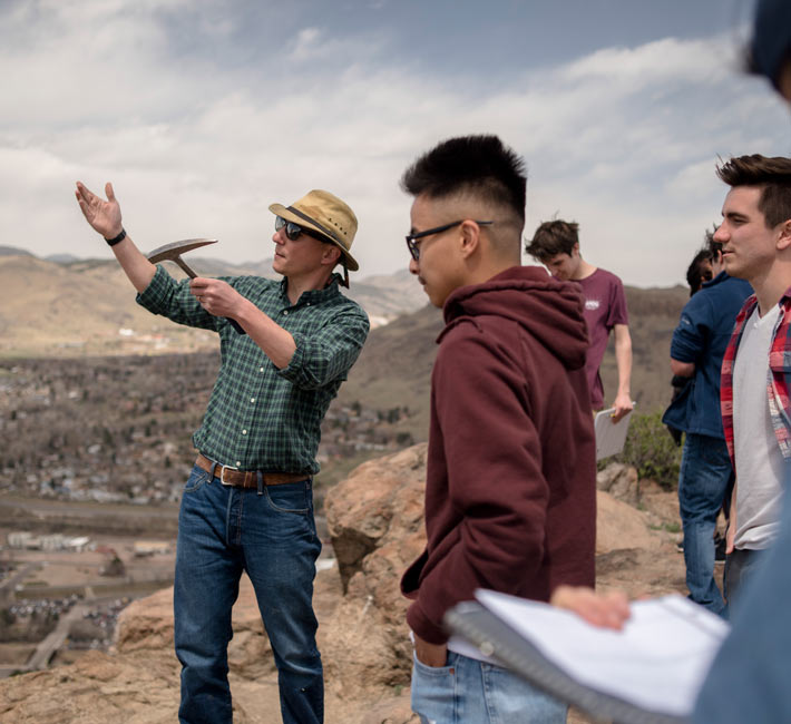 Instructor teaching students with a rock hammer during a ecology field study overlooking a landscape.
