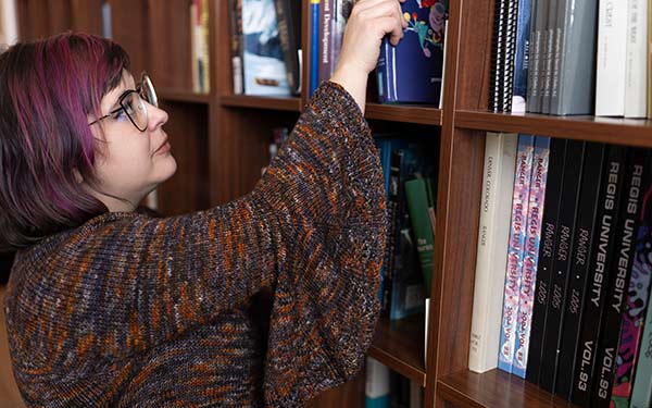 Person with purple hair reaching for a book on a tall wooden bookshelf filled with books.