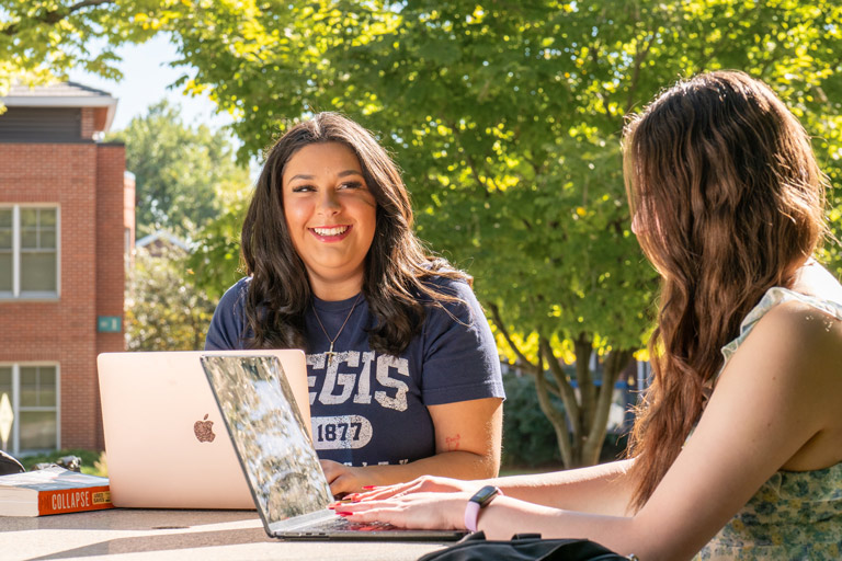 Two college students work together on laptops at a sunny campus table, smiling during a friendly outdoor study session near a brick building