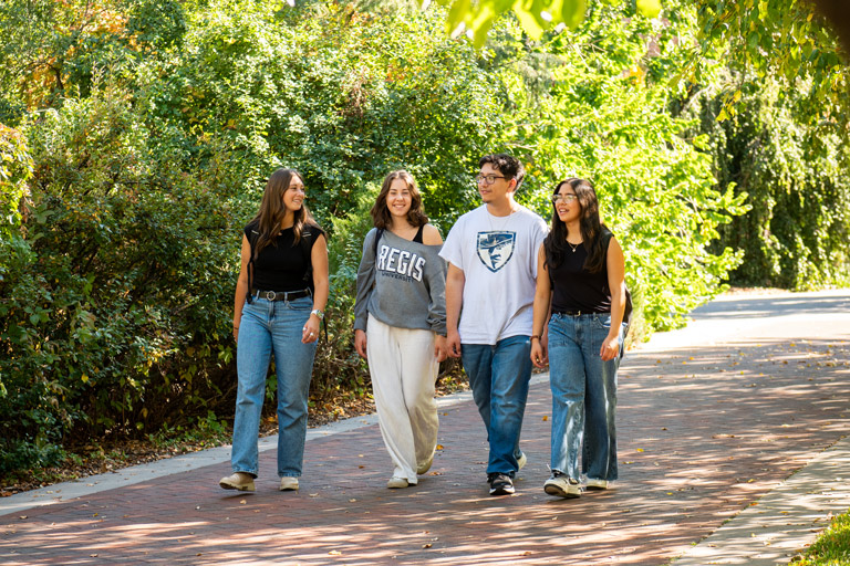 Four students walking across campus in the fall with green foliage and yellowing trees in the background
