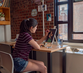 Student seated at her desk in her dorm, painting a canvas