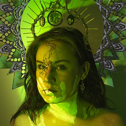 photo of a woman wearing an elaborate headpiece with a paint handprint on her face and bathed in green light