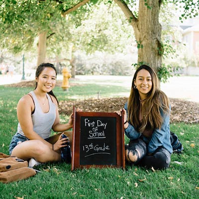 Two young women sit in the grass beside a chalkboard sign that reads "First day of 13th grade"