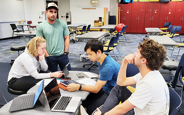 Students in the Regis Manual Physical Therapy Fellowship Certificate sit or stand in a classroom working on their laptops on a group project