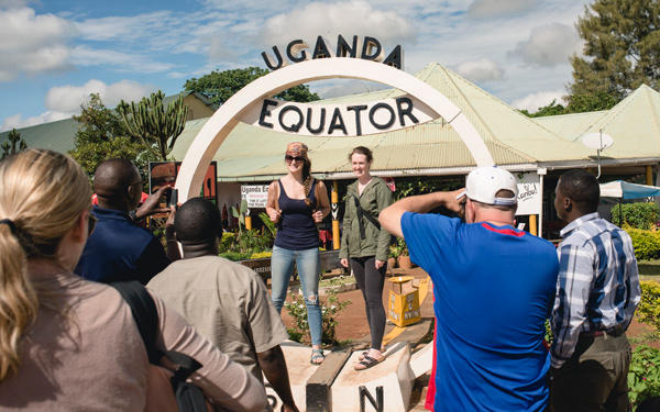 Two Regis students stand under a large arch labeled 'Uganda Equator' as a group gathers to take photos. A small marketplace is visible behind them.