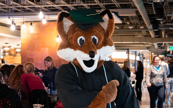 Regi the mascot in graduation attire interacting with students in a campus gathering space