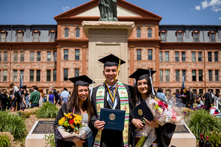 three graduates wearing regalia proudly display their diplomas and bouquets of flowers in front of Main Hall on the Quad