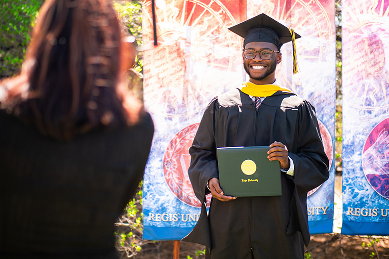 a graduate wearing cap and gown poses for a photo with his diploma at a commencement ceremony on the Regis campus