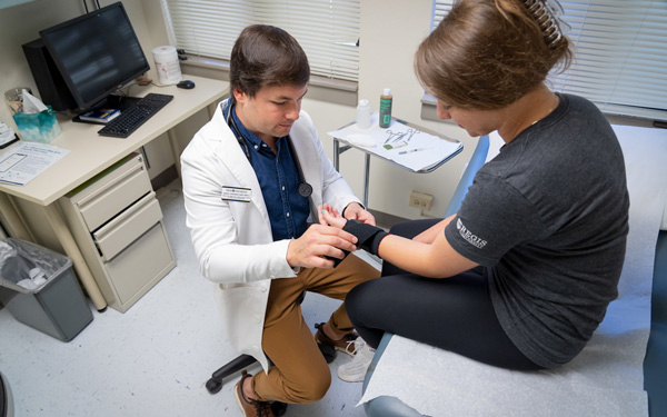 A healthcare professional in a white coat assists a patient with a wrist brace in a medical exam room at Regis University