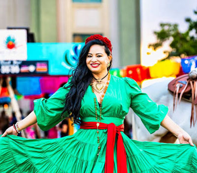 Smiling woman in a bright green dress with a red sash spreads her skirt at an outdoor fair, with colorful stalls and a saddled horse behind her