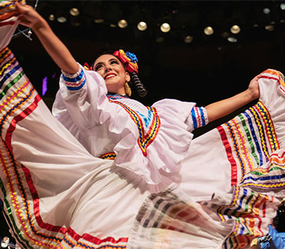 Smiling dancer in a white, vividly embroidered dress twirls onstage, arms raised and skirt flowing in a joyful cultural performance.