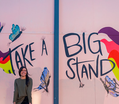 woman standing in front of a colorful mural, "TAKE A BIG STAND" written on the wall