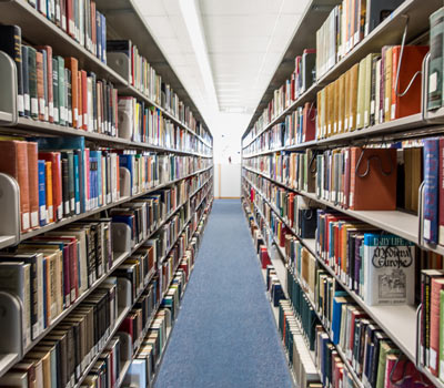 A quiet library aisle stretches between tall shelves filled with colorful books, creating a spacious, orderly, and inviting atmosphere.