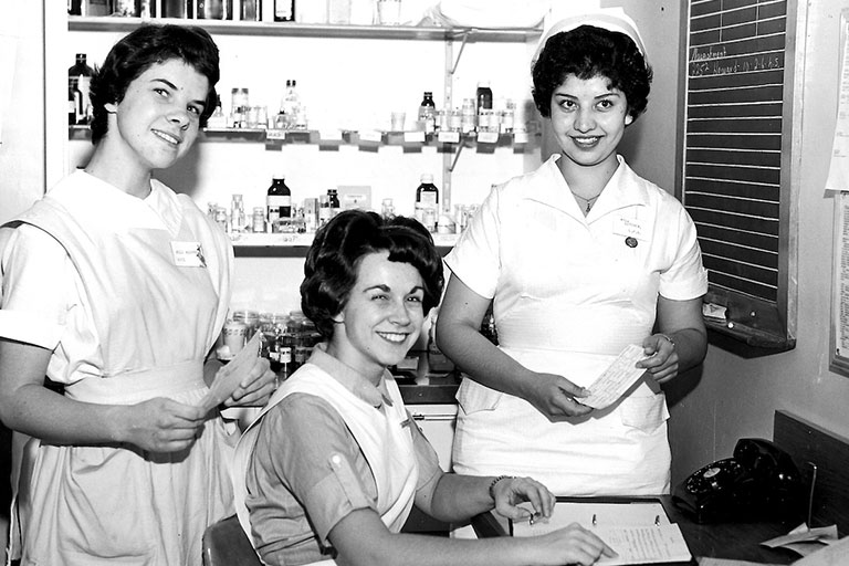 historical black and white photo of nurses in old-fashioned nursing uniforms working and smiling for the camera