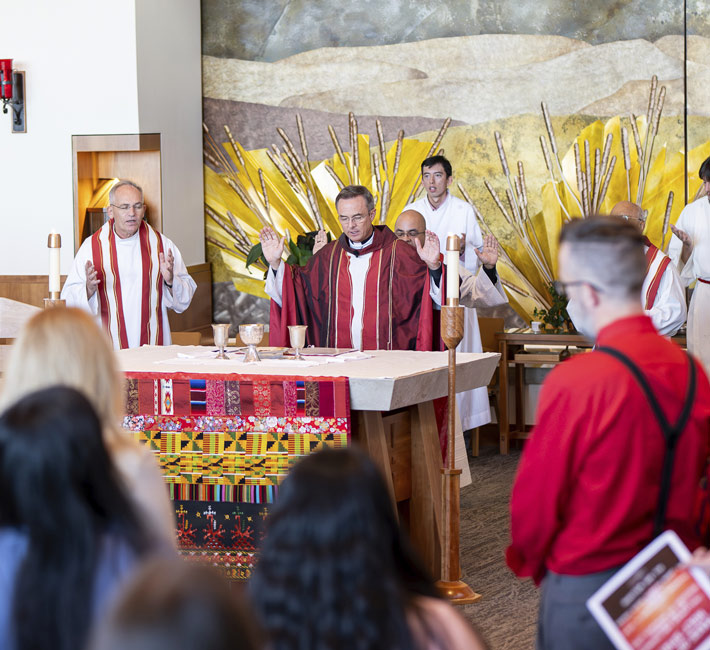 Fr. Hendrickson stands at an altar by two servers, facing a seated congregation in a St. John Francis Chapel. The background features a large, textured landscape mural and a blue religious tapestry under a vaulted ceiling.