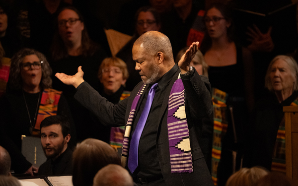 Roger Holland, wearing a purple tie and stole, conducts a choir in performance, with choir members singing in the background