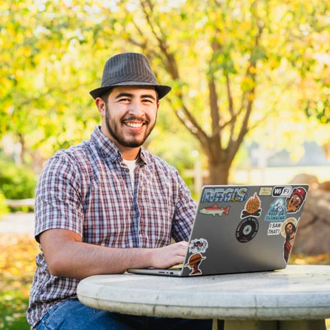 Student sitting at an outdoor table using a laptop with trees in the background
